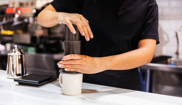 Método de preparo do café com sistema Aero Press. Mulher barista de camiseta preta pressiona Aeropress para encher um copo com café expresso no balcão de mármore. Café profissional para fazer café.