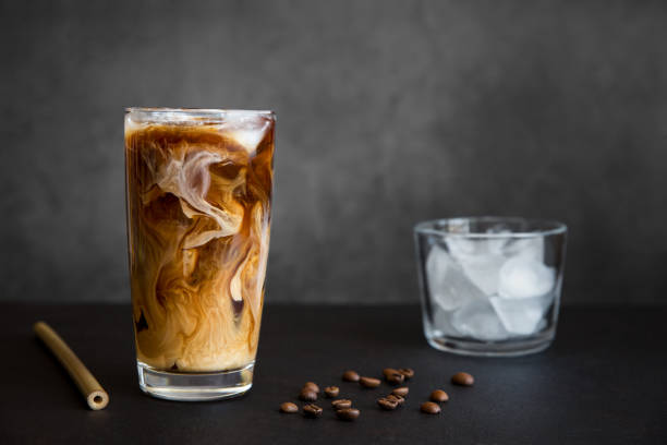 Café gelado em copo alto com creme, recipiente com gelo, palha de coquetel e grãos de café em fundo escuro com espaço de cópia. Bebida refrescante.