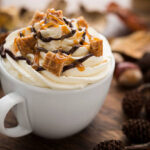 Bebida café cremoso (chocolate quente ou Café Aromatizado) com Chantilly, pedaços de Waffle de Caramelo, calda de caramelo e calda de Chocolate, rodeada de Folhas de Outono secas.