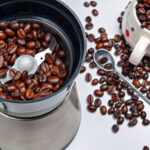Moedor de café elétrico com grãos de café torrados dentro e grãos de café espalhados sobre uma mesa com uma colher de metal de cima e uma xícara cheia de grãos de café, vista de perto de cima