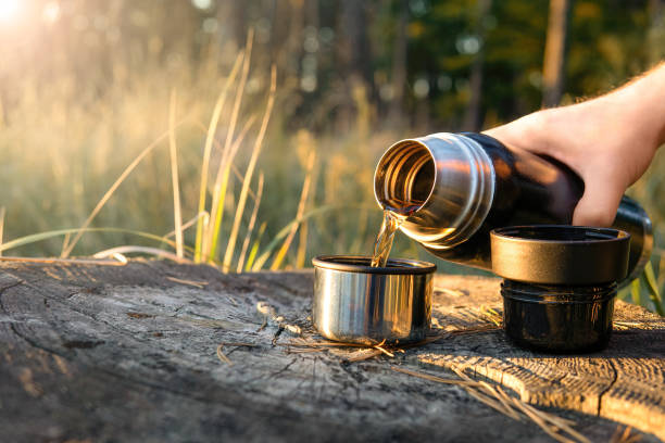 Turista derramando café quente de uma garrafa térmica em duas xícaras em pé no tronco de uma árvore ao pôr do sol. Conceito de caminhadas, viagens e estilo de vida ativo